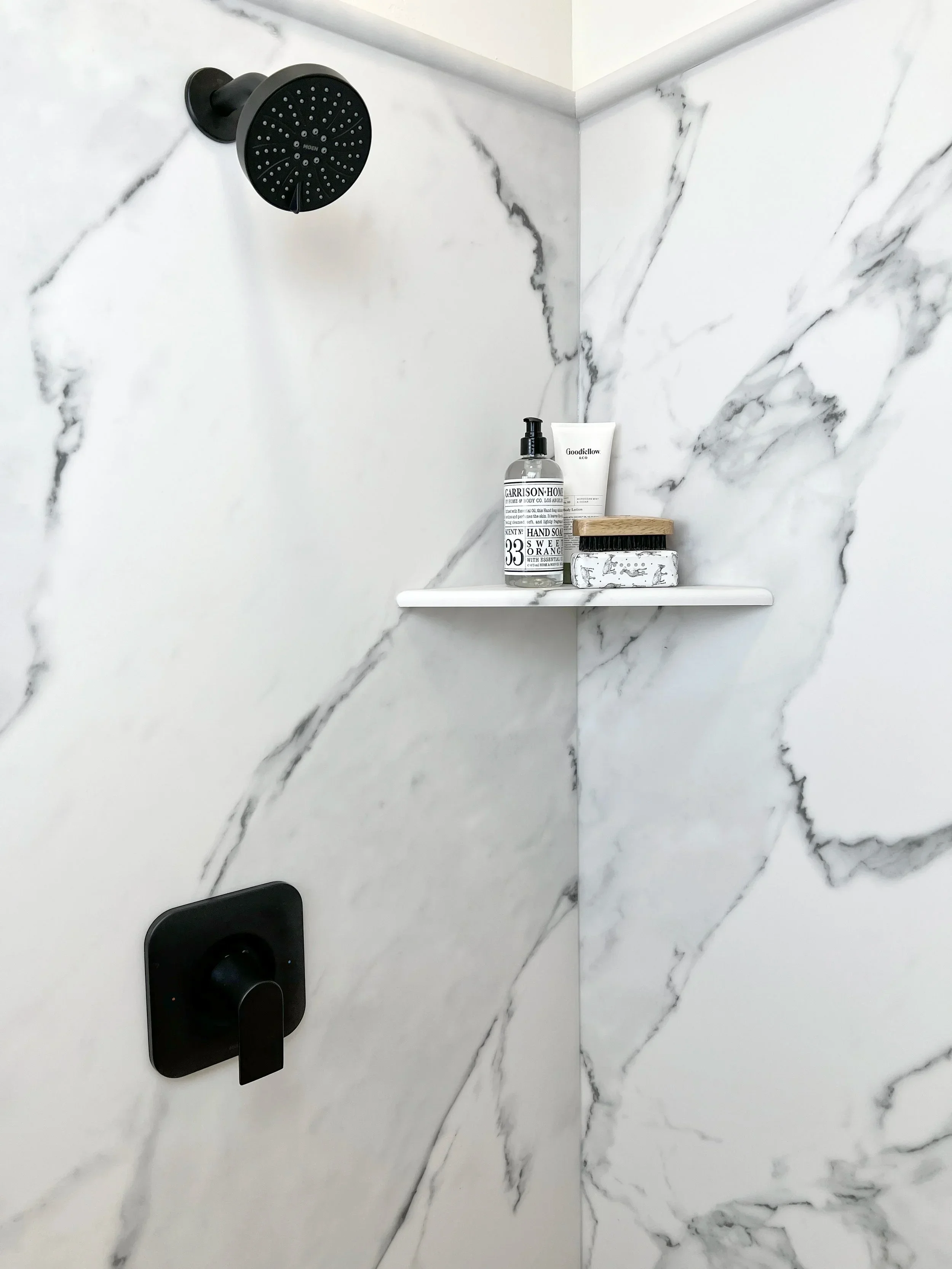 Marble tile shower with matte black rainfall showerhead and valve trim, floating shelf with bath products