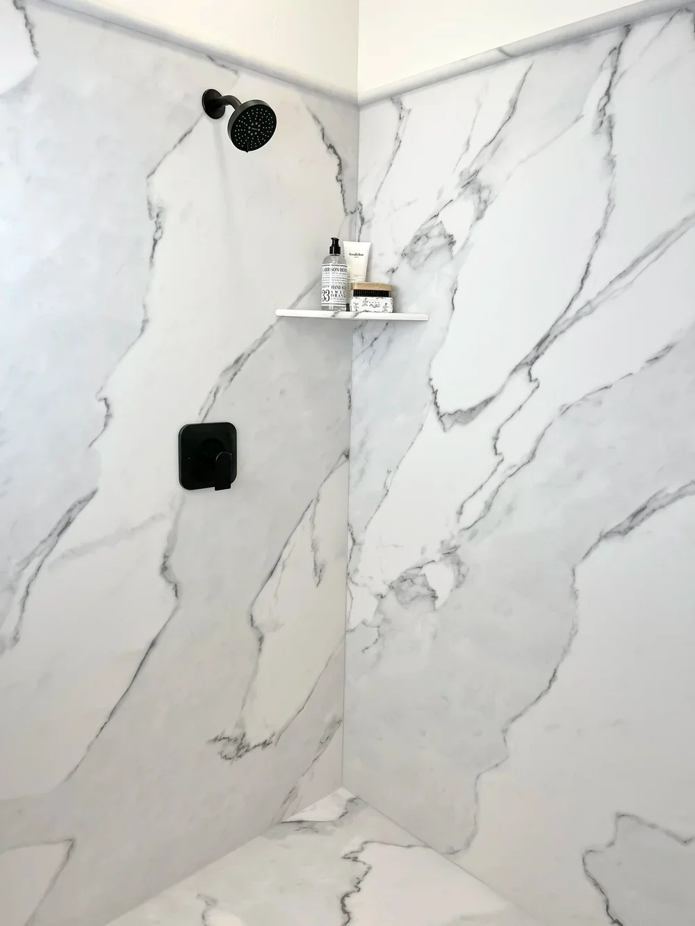 White marble-look tile shower with black rainfall showerhead and floating shelf