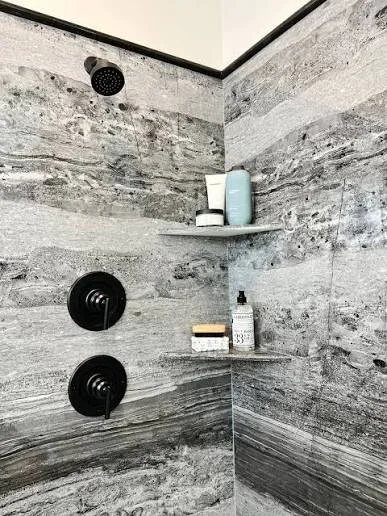 Shower with gray stone-look tile, black fixtures, rainfall head, and floating shelf