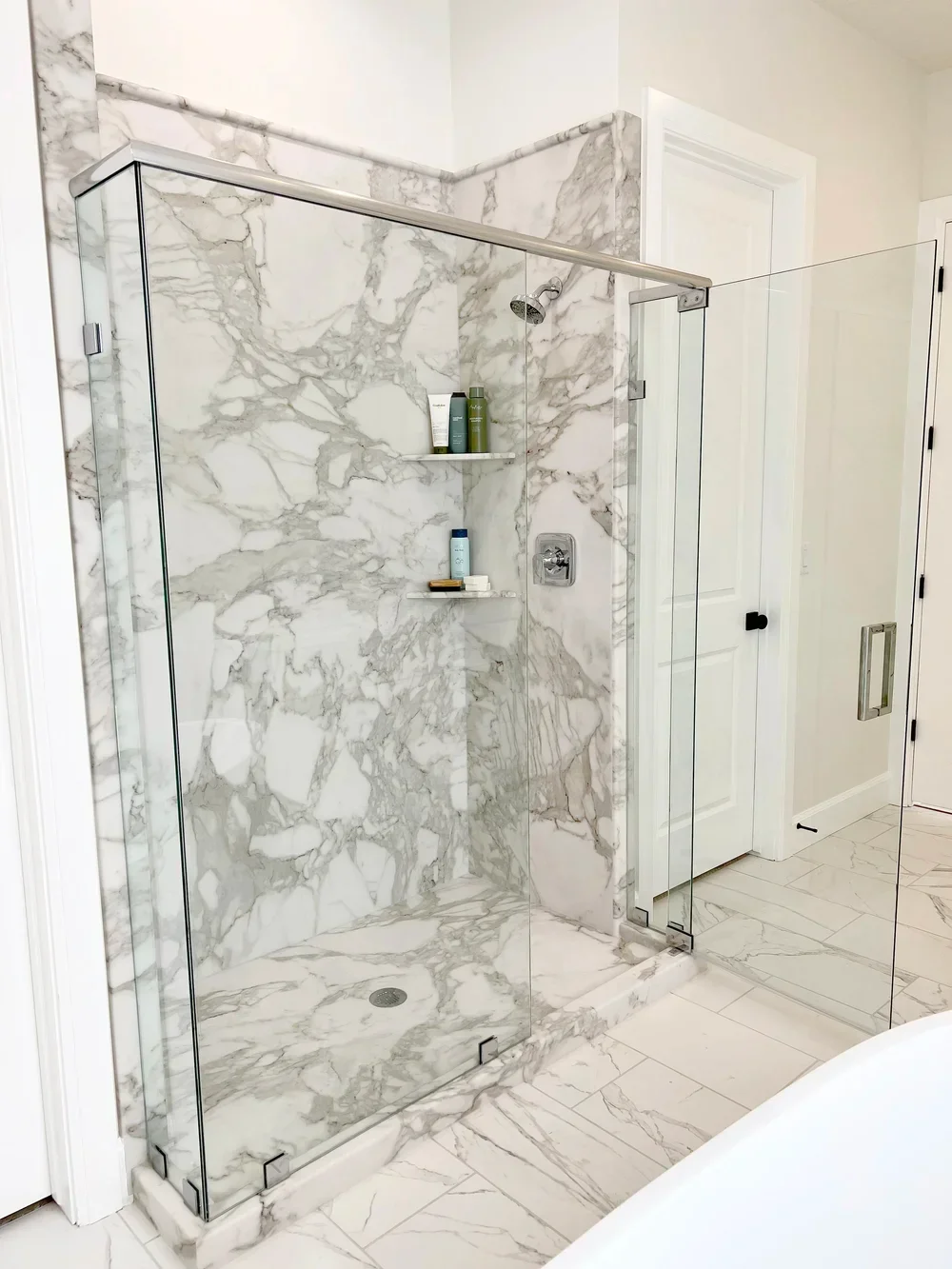 Frameless glass shower with Calcutta marble tile walls, floating shelves, and chrome fixtures